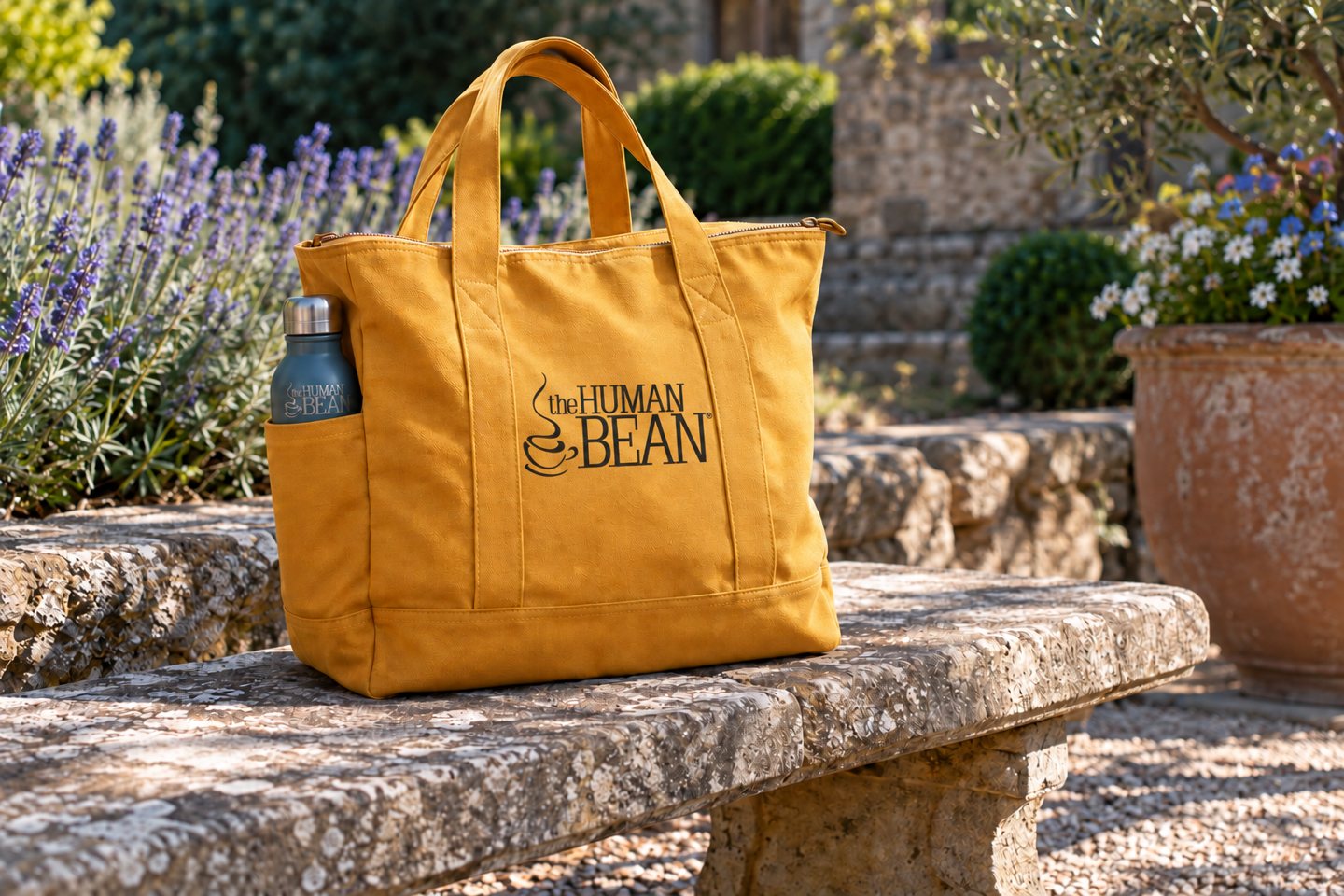 An ochre zipper tote with a printed logo and a branded reusable bottle on a garden bench.